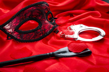 Concept image with a mask, hand cuffs and a flogger covered in rose petals on red silk. BDSM is a...
