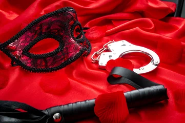 Concept image with a mask, hand cuffs and a flogger covered in rose petals on red silk. BDSM is a variety of erotic practices or role playing involving bondage, dominance and submission, masochism © Victor Moussa