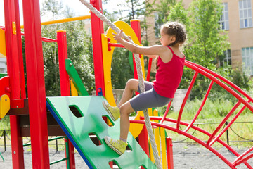 Little caucasian girl dressed in a red jersey playing on playground, climbing the wall on a rope