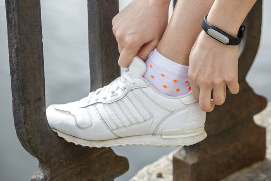 Woman hands with black fitness bracelet adjusts laces on white sneakers. On the street, on the parapet of the river.