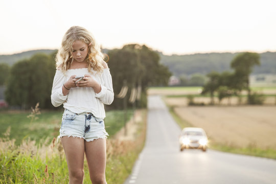 Sweden, Blekinge, Solvesborg, Teenage girl (14-15) with mobile phone standing at side of road