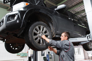 Mechanic diagnosing car wheel with screwdriver. Car maintenance in garage. Young auto service worker fixing wheel on raised on hoist car.