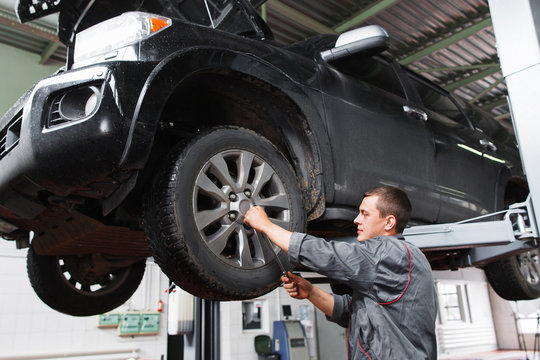 Mechanic Diagnosing Car Wheel With Screwdriver. Car Maintenance In Garage. Young Auto Service Worker Fixing Wheel On Raised On Hoist Car.