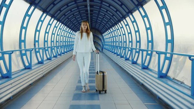 Young business woman with travel bag on wheels is on the transition from the terminal at the airport or train station