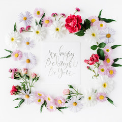 inspirational quote "be happy, be bright, be you" written in calligraphy style on paper with pink, red roses, chamomiles and leaves isolated on white background. Flat lay, top view