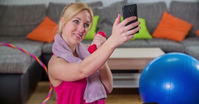 A lady is taking a few selfies after a successful work out. She looks happy. She's holding a light weight in her left hand. Wide-angle shot.
