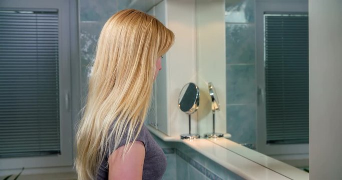 A young lady is standing in front of a mirror in the bathroom. She is taking a toothbrush and a toothpaste and is about to start cleaning her teeth.
