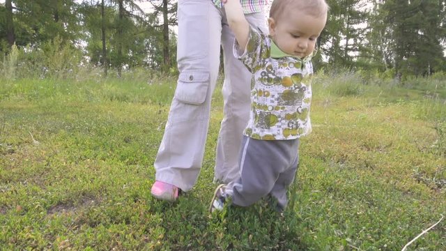 Mum learns  child walk outdoors. Baby first steps on grass, slow motion.