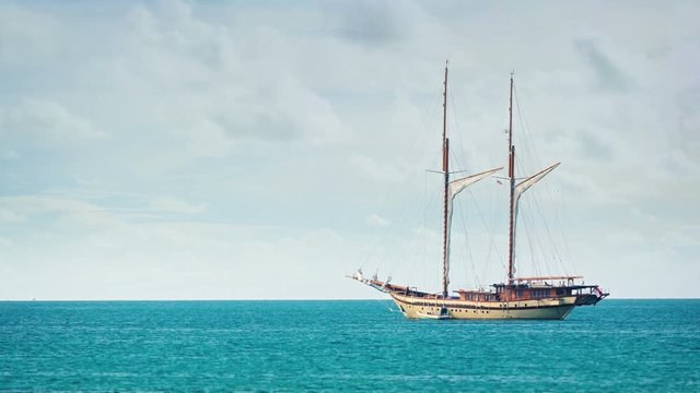 Two masted, wooden sailing yacht in extreme zoom, anchored on the horizon off a popular tropical beach in Southeast Asia. Video 3840x2160