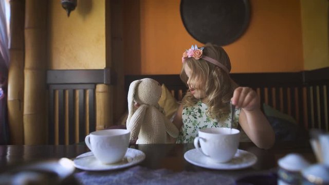 A little girl is having tea with her stuffed rabbit in a cozy cafe.