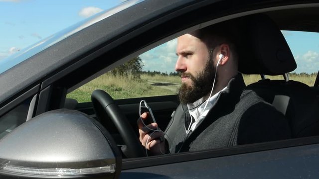 young handsome hipster man sits in the car and listens music on smartphone - countryside - sunny weather 