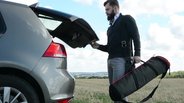 young handsome hipster man goes to car trunk and load trunk with bags - countryside