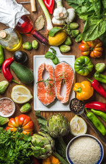 Dinner cooking ingredients. Raw uncooked salmon with vegetables, rice, herbs, lemon, artichokes, spices and bottle of rose wine on white ceramic board over rustic wooden background, top view