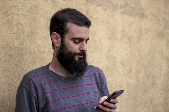 Young man with beard checking smartphone and wall of a house in