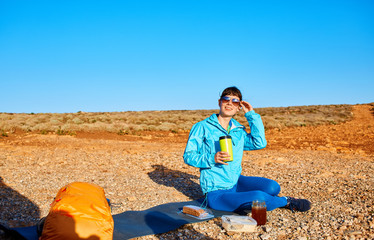 woman traveler sitting on the rug near the car and drink coffee. a woman in a place like a desert at dawn