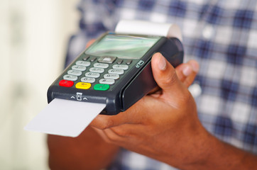 Man wearing blue white square pattern shirt holding up credit card payment terminal in front of camera, closeup angle