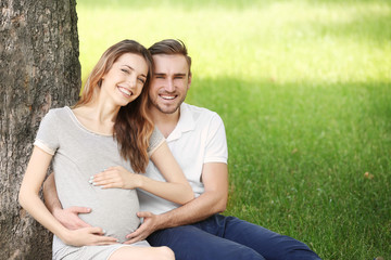 Happy pregnant woman and her husband  sitting at tree in park