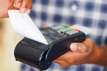 Man wearing blue white square pattern shirt processing payment using credit card terminal in front of camera, closeup angle