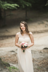 Fototapeta na wymiar Gorgeous bride in elegant dress holding bouquet posing near forest and lake