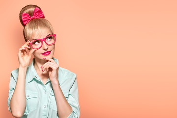 Fashion. Woman in Stylish Glasses Having Fun. Hipster fashion girl think, idea. Playful nerd Blonde with Glamour Pinup Stylish hairstyle, Trendy fashion, red bow Makeup. Unusual Creative, on yellow