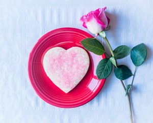 Pink heart sugar cookie on red plate next to a pink rose on white textured background.