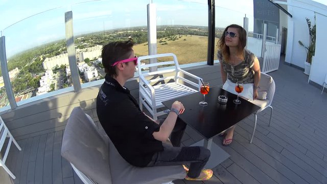 Lesbian couple date, girls sits on terrace in rooftop bar in luxury hotel restaurant, smile, laugh, drink cocktails and clink glasses on summer vacation, Spain, Majorca