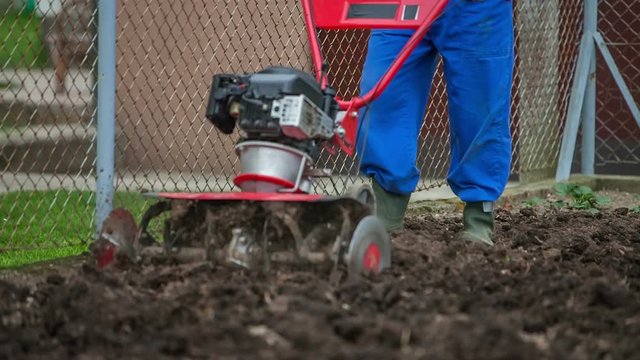 A farmer is ploughing a garden with the help of a cultivator in early spring. The machine is helping him to turn the soil over. Wide-angle shot.
