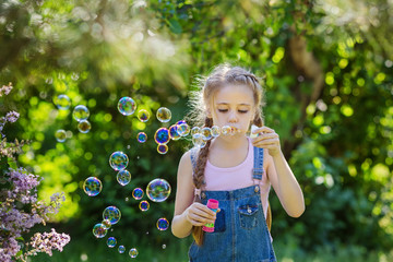 Portrait of beautiful little girl in denim overalls blowing bubbles in lilac blossoming garden