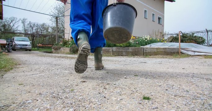 A senior farmer is walking back from the field to the front yard of his house. He was sowing wheat and is now having an empty bucket in his hand. Close-up shot.
