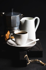Italian coffee set for breakfast. Cup of hot espresso, creamer with milk, cantucci and moka pot on dark rustic wooden board over black background