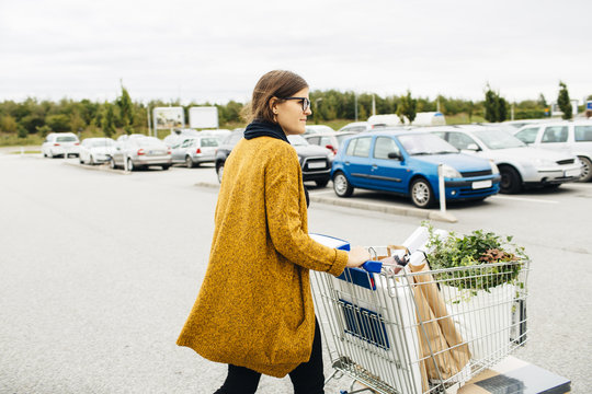 Side view of young woman pushing trolley full of purchases towards car park