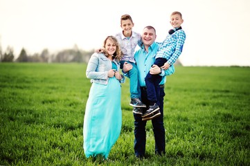 Happy pregnant family with two sons, dressed in a turquoise clothes, spending time together at green grass at field