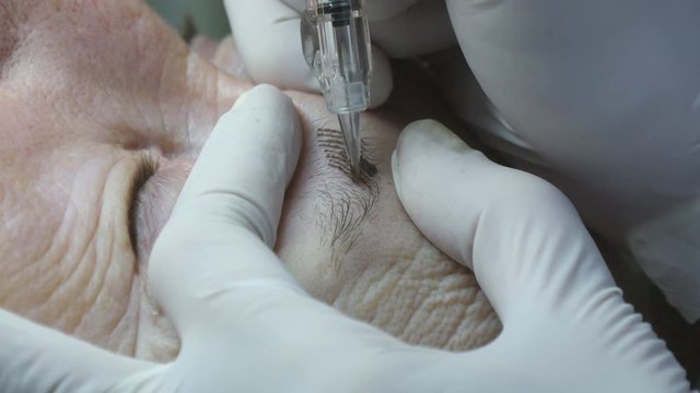 Old woman lies and getting eyebrows makeup at beauty salon. Applying Permanent Make up on eyelash. Beautician making permanent makeup of eyelash. Master works with the eyebrows. Semi-permanent make-up