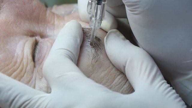 Old woman lies and getting eyebrows makeup at beauty salon. Applying Permanent Make up on eyelash. Beautician making permanent makeup of eyelash. Master works with the eyebrows. Semi-permanent make-up