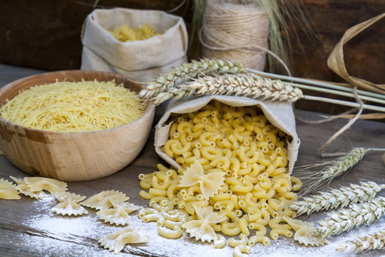 Uncooked Italian pasta Farfalle, Elbow macaroni and Fusilli in canvas little bag, scattered flour with wheat ears on dark wooden background. Shallow depth of field. Shallow depth of field