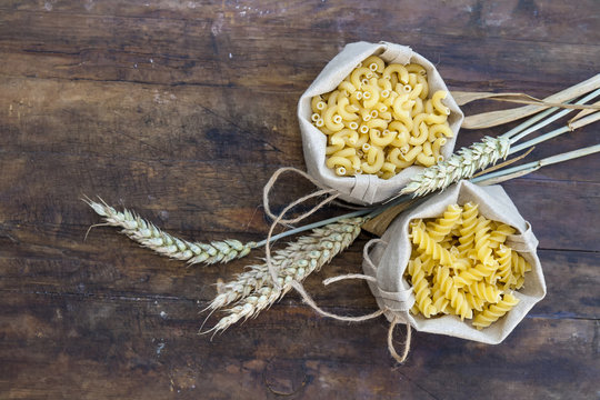 Uncooked Italian pasta Elbow macaroni and Fusilli in canvas little bag with wheat ears on dark wooden background. Shallow depth of field