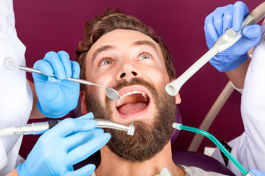 Close up of men patient with beard with open mouth and white straight teeth sitting in medical chair. Doctor with assistant making check up in dental clinic