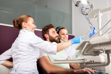 Female doctor and her smiling assistant showing teeth x-ray to male patient in dental hospital. Handsome men with beard sitting in medical chair and looking at the picture.