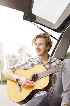 Portrait of smiling young man sitting at backside of his car playing guitar