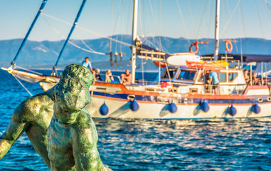Obraz premium Daily trip on Island of Brac / .Sculpture of fisherman in foreground with touristic boat in background in Island of Brac, Bol town, Croatia.