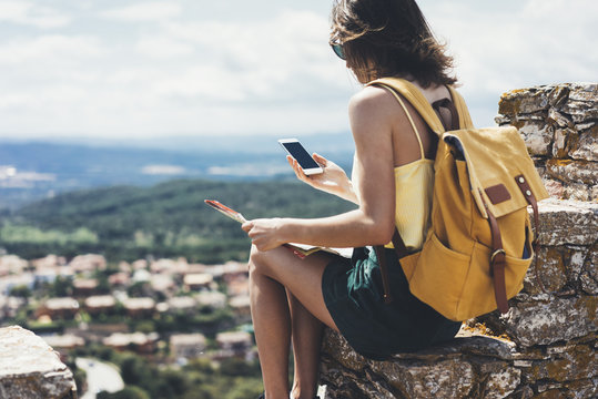 Hipster young girl with bright backpack looking a map. View from the back of the tourist traveler on background mountain, sea. Mock up for text message. Female hands using smartphone, holding gadget
