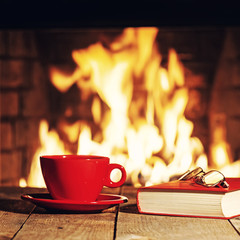 Red cup, glasses and old book near fireplace on wooden table.