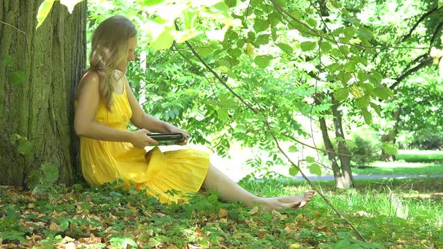 Beautiful woman girl in dress sit near tree trunk and start using laptop computer in park. 4K