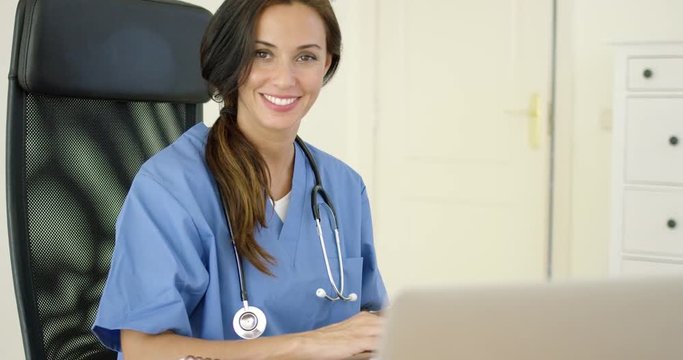 Beautiful female doctor with stethoscope grinning while typing on laptop computer at desk with black leather chair in office