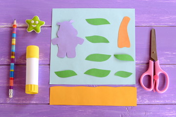 Hippo, leaves, palm tree trunk cut from colored paper. Set for children craft projects. Paper card hippo craft. Glue stick, scissors, pencil, eraser on wooden background. Top view