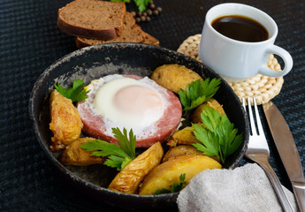 Fried egg in salami and baked potatoes. Submission of a frying pan with a cup of coffee and rye bread. Breakfast.