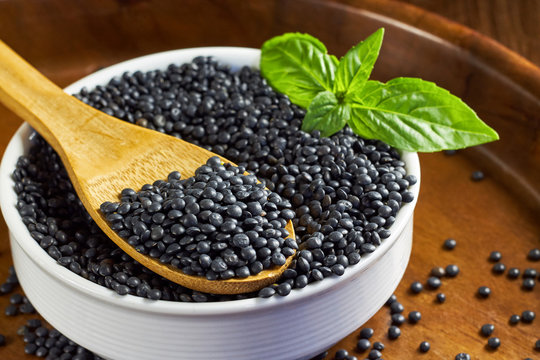 Round white bowl full of beluga lentils and basil leaves with wooden spoon on brown wooden background.