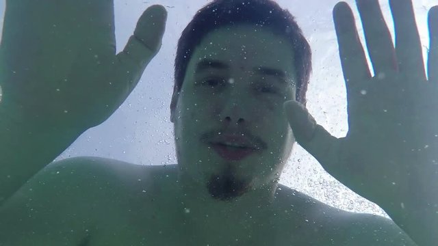 A young guy is waving to the camera when he is driving under water. He then passes it and continues swimming. Close-up shot.
