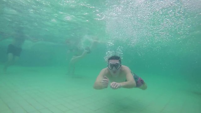 A young man is having his googles on and he is swimming under surface. He gives thumbs up and smiles into the camera.

