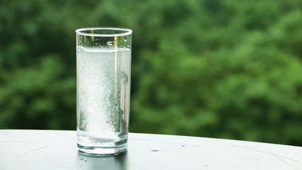 White soluble tablet (meltlets) dissolving in a water glass against green nature background (trees under wind and rain)
